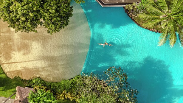 An aerial view of a resort with a clear blue pool, lush greenery, and a sandy beach area.
