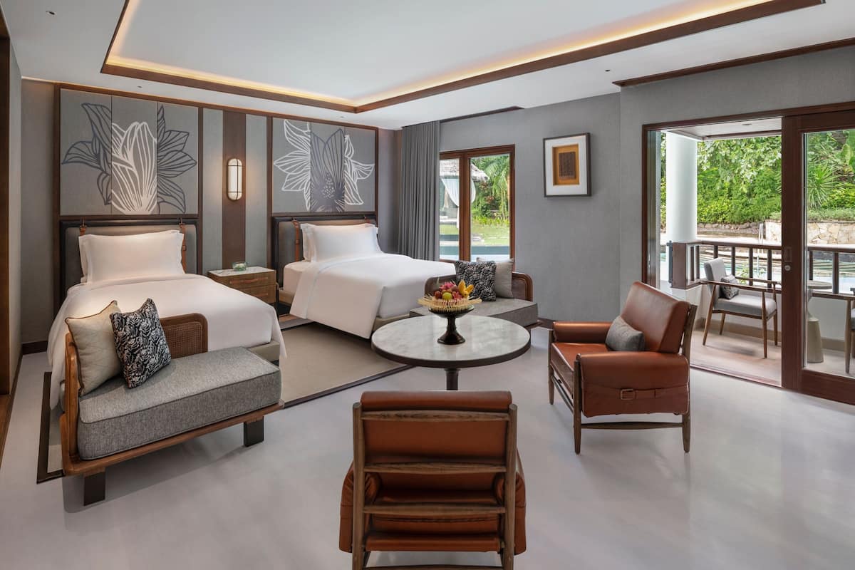 A modern hotel room with a large bed, a sofa, a chair, a small table with fruit, and a balcony with a view of greenery.