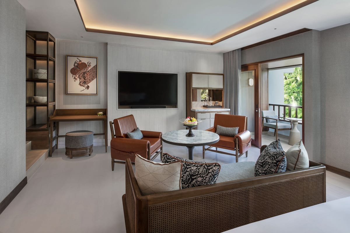 A modern living room with a sofa set, a round coffee table, and a flat-screen TV mounted on the wall. There is a balcony with a view of greenery.