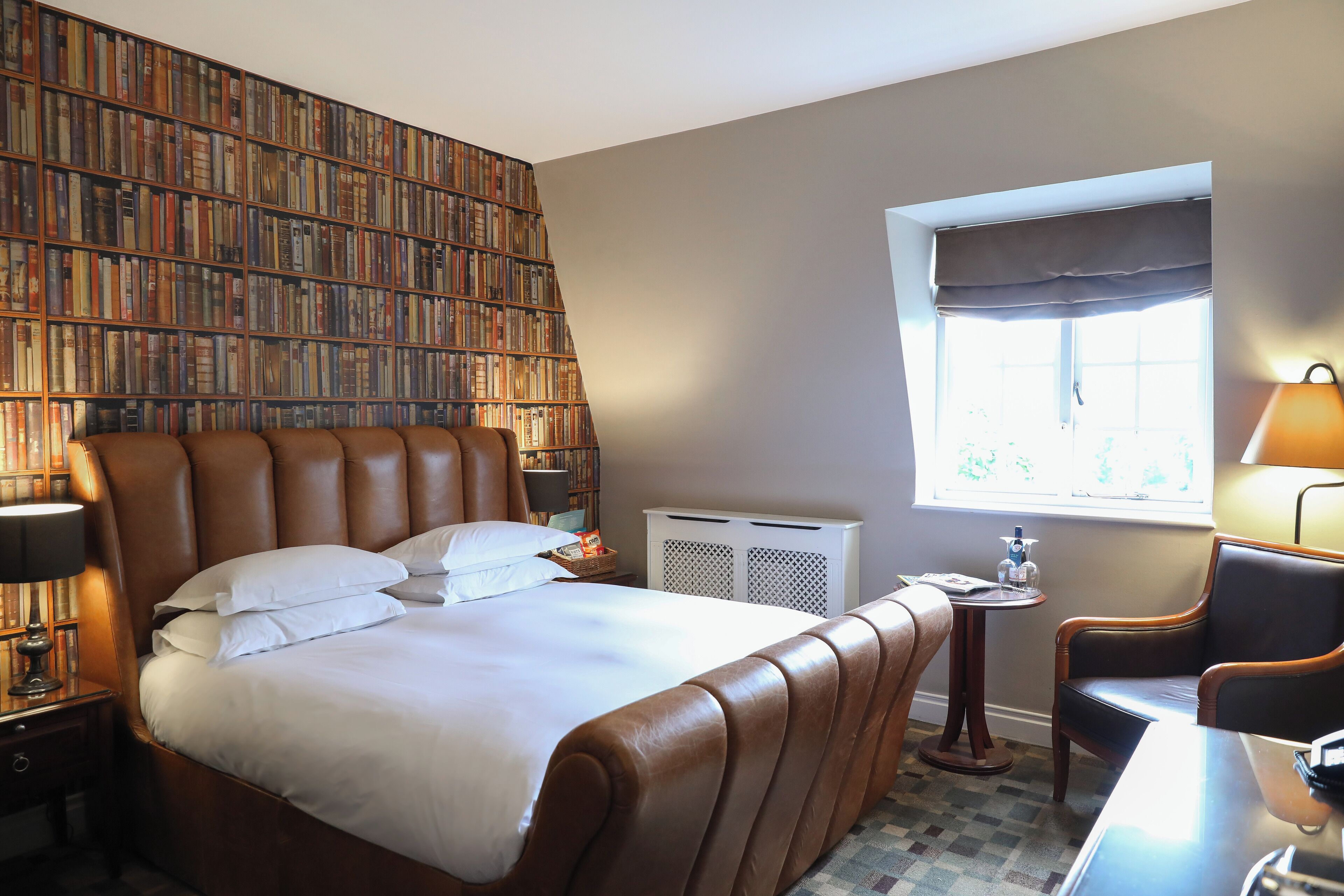 Une chambre d’hôtel avec une grande bibliothèque, un lit doté d’une tête de lit en cuir marron, une table de chevet avec une lampe et une petite table ronde avec une chaise.