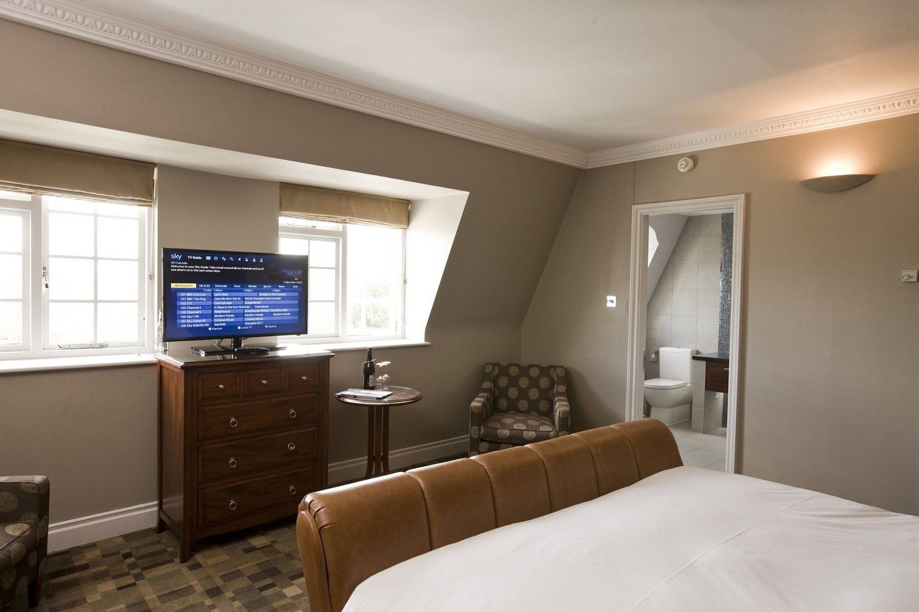 A hotel room with a bed, a TV on a dresser, a chair, and a bathroom visible through an open door.