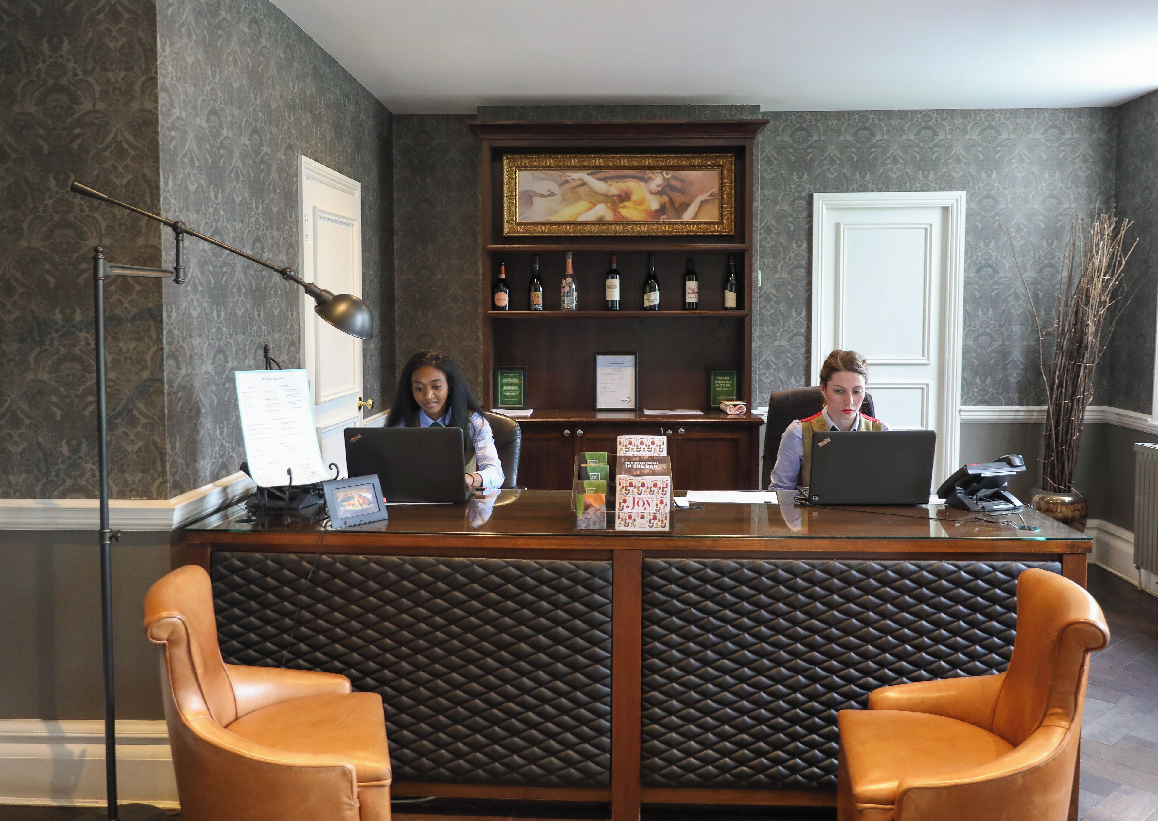 A reception desk with two laptops, a framed painting above, and a shelf with bottles.