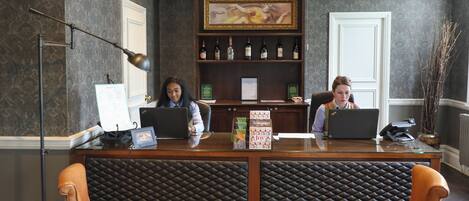 A reception desk with two laptops, a framed painting above, and a shelf with bottles.