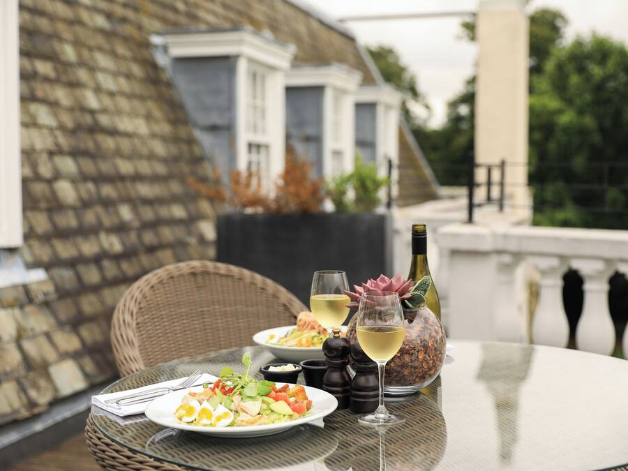Una mesa de comedor con platos de comida, dos copas de vino blanco y una botella de vino en un balcón con sillas de mimbre y un muro de piedra al fondo.