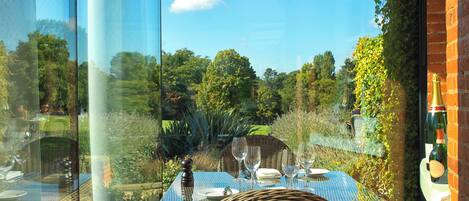 A dining table set for two with wicker chairs, a glass table, and a view of a garden through a glass partition.