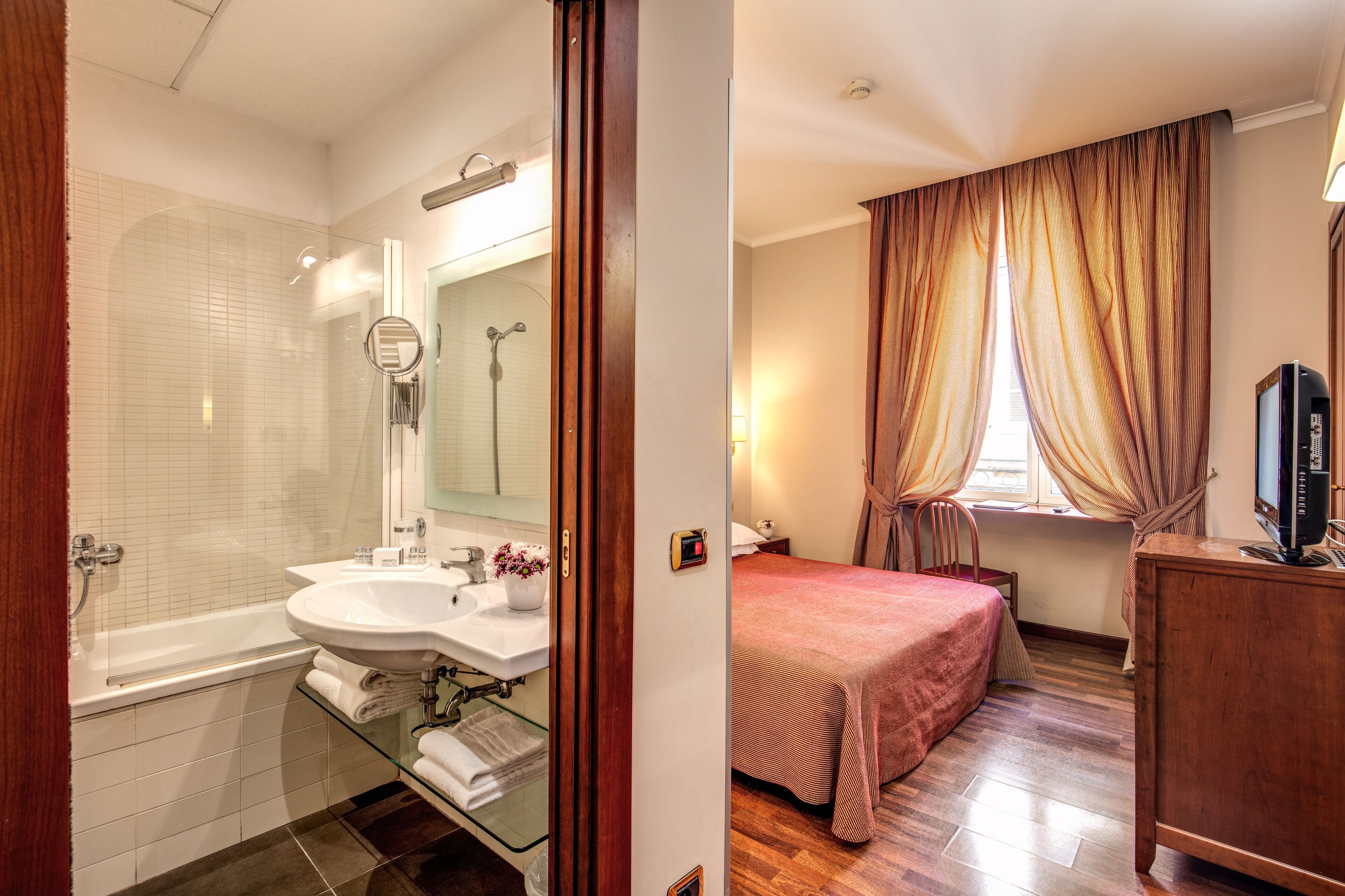 A bathroom with a bathtub, sink, and mirror. The room has wooden flooring and a bed with a red bedspread.