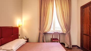 Un letto ben rifatto con testiera rossa, una sedia in legno, un comodino con una lampada e una finestra con tende.