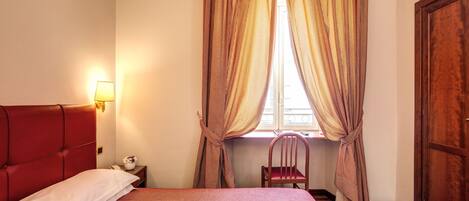 Una cama bien hecha con cabecera roja, una silla de madera, una mesita de noche con una lámpara y una ventana con cortinas.
