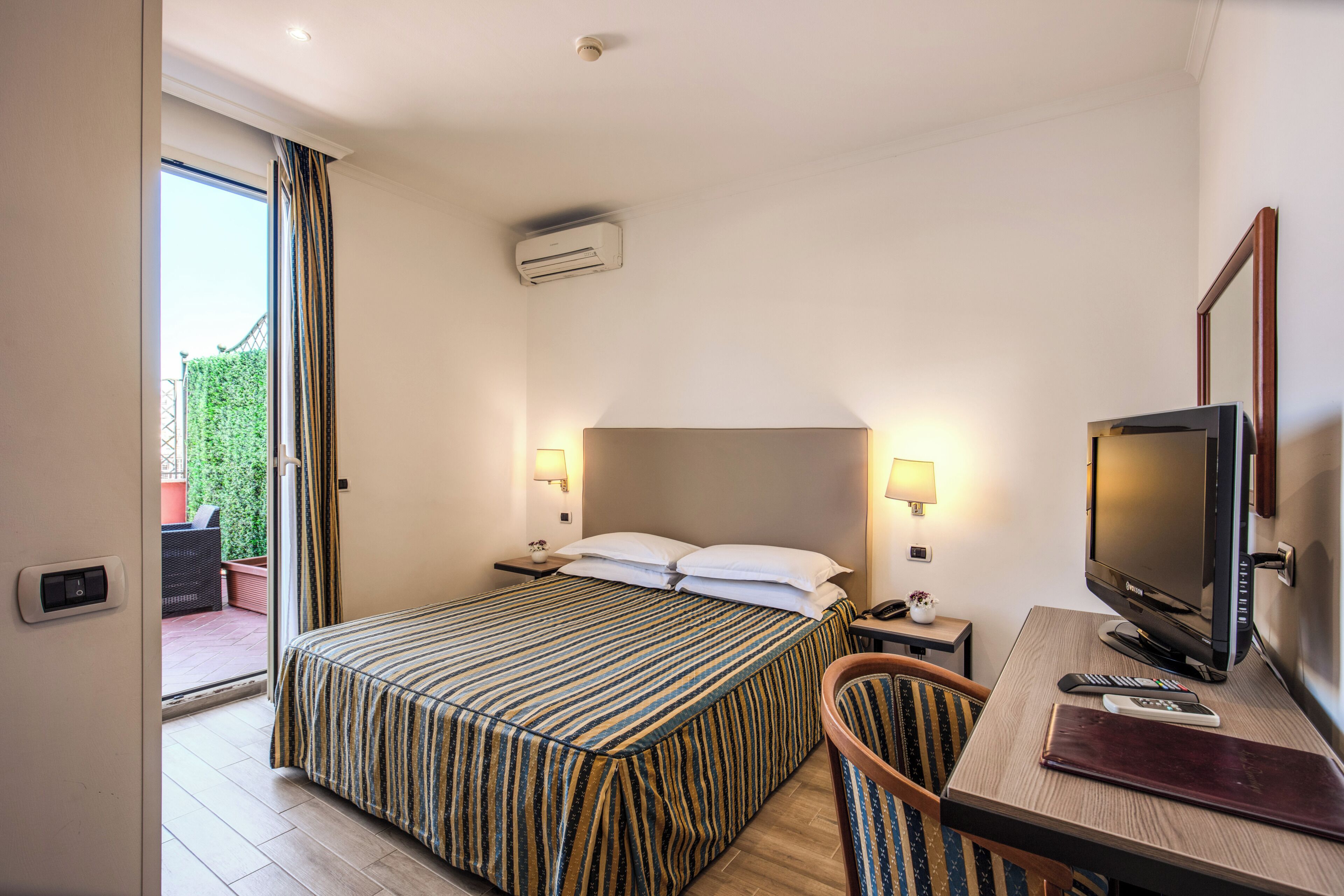 A hotel room with a bed, a desk with a TV, a chair, and a window with curtains.