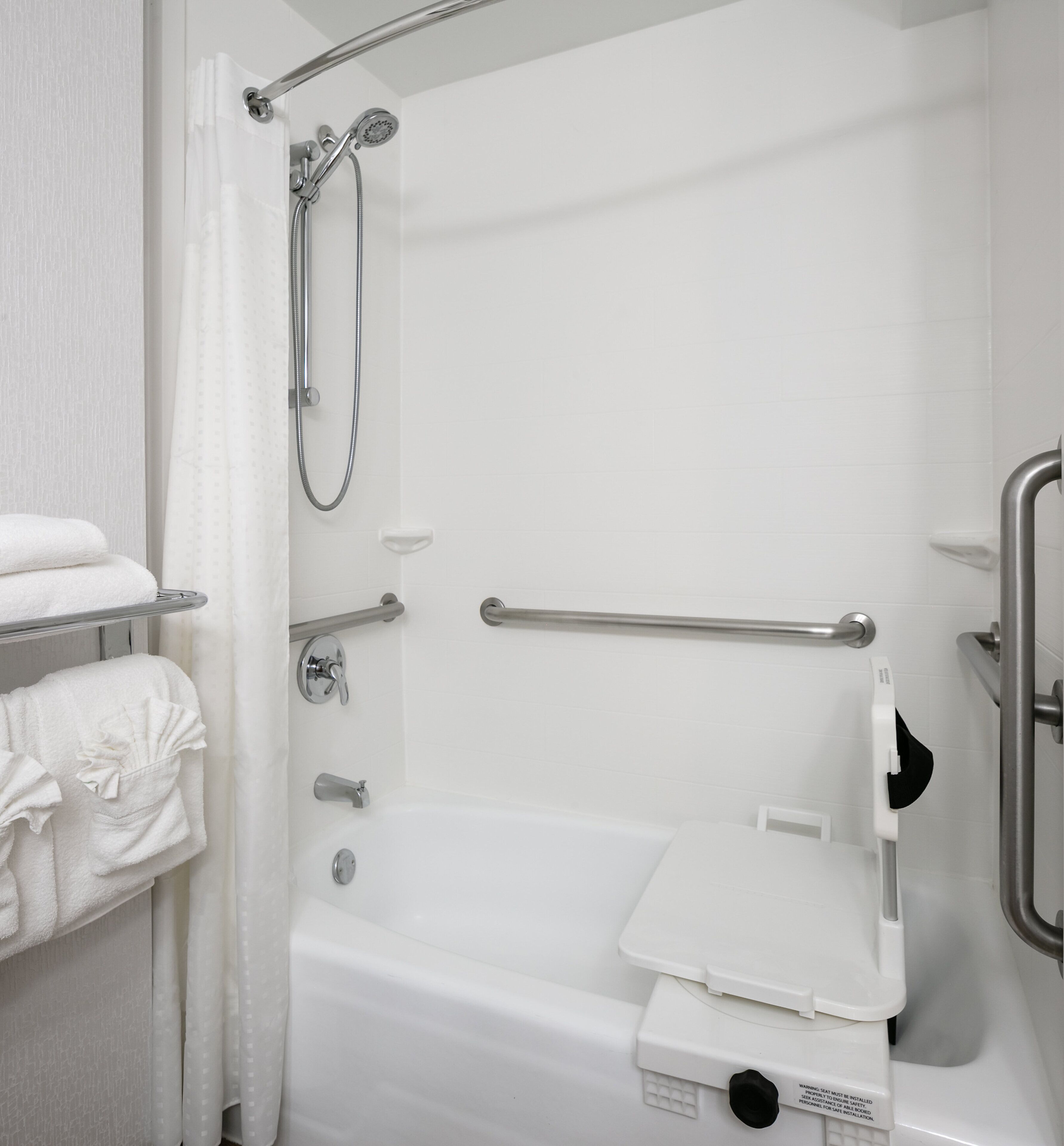 Une salle de bain équipée d’une douche, de barres d’appui, de toilettes avec un siège de sécurité et de serviettes rangées sur un porte-serviettes.