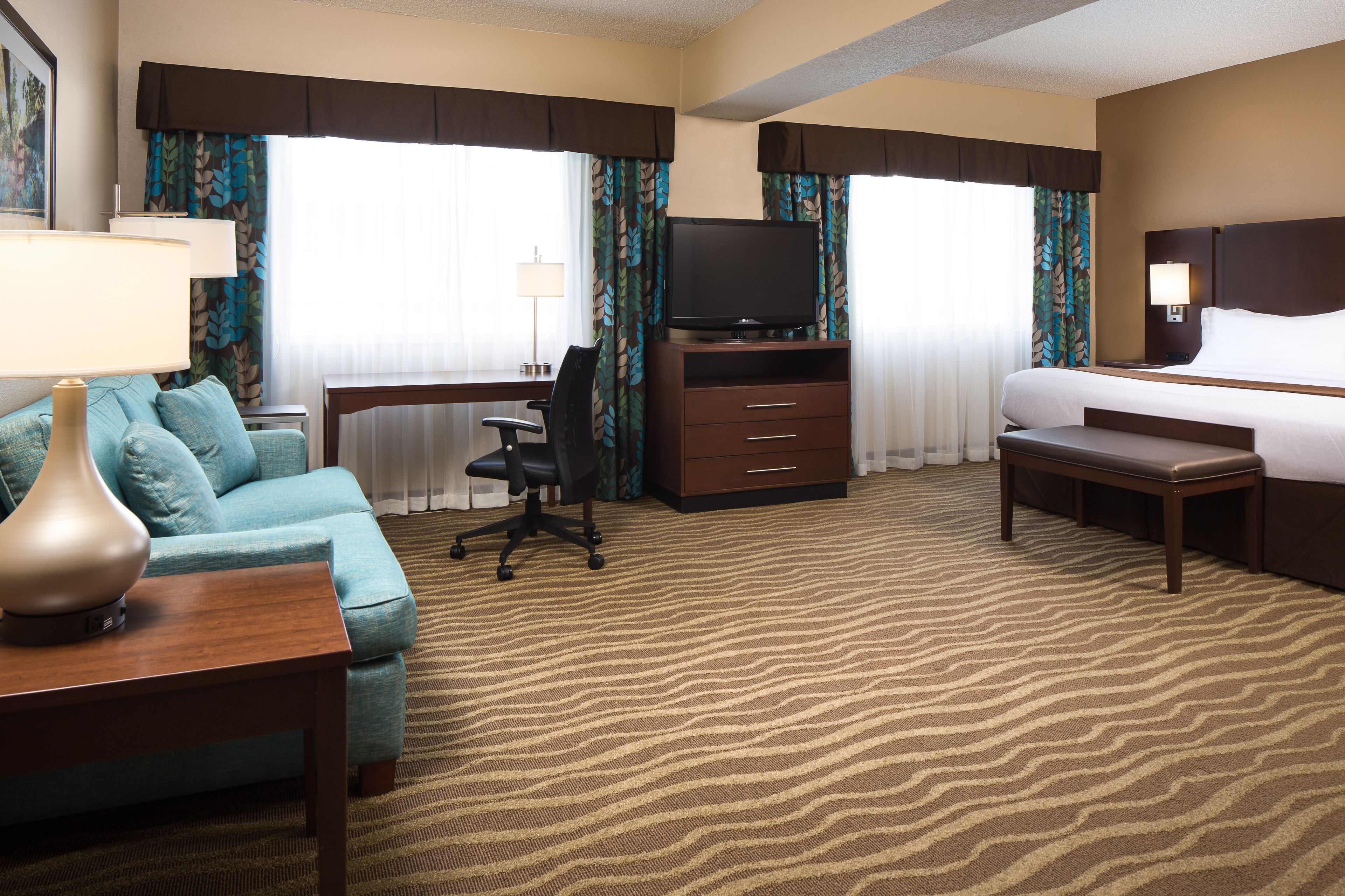 A hotel room with a sofa, a desk with a lamp, a television, a bed, a bench, and a carpeted floor.