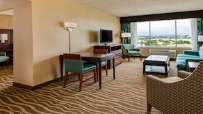 A hotel room with a desk, television, two chairs, a sofa, and a large window offering a city view.
