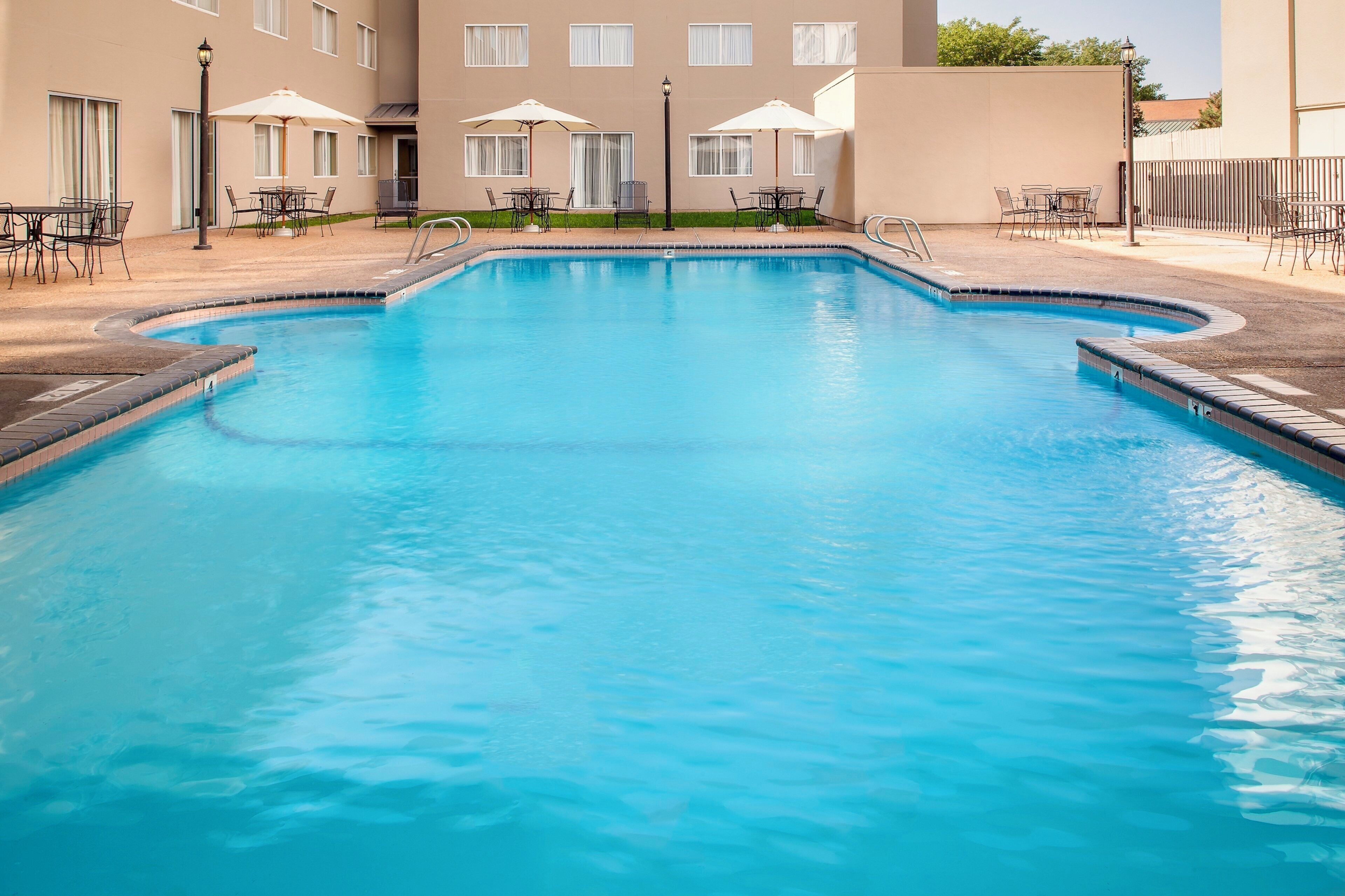 A rectangular swimming pool with a tiled edge, surrounded by a low brick wall. There are outdoor tables and chairs with umbrellas, and a building with multiple windows in the background.