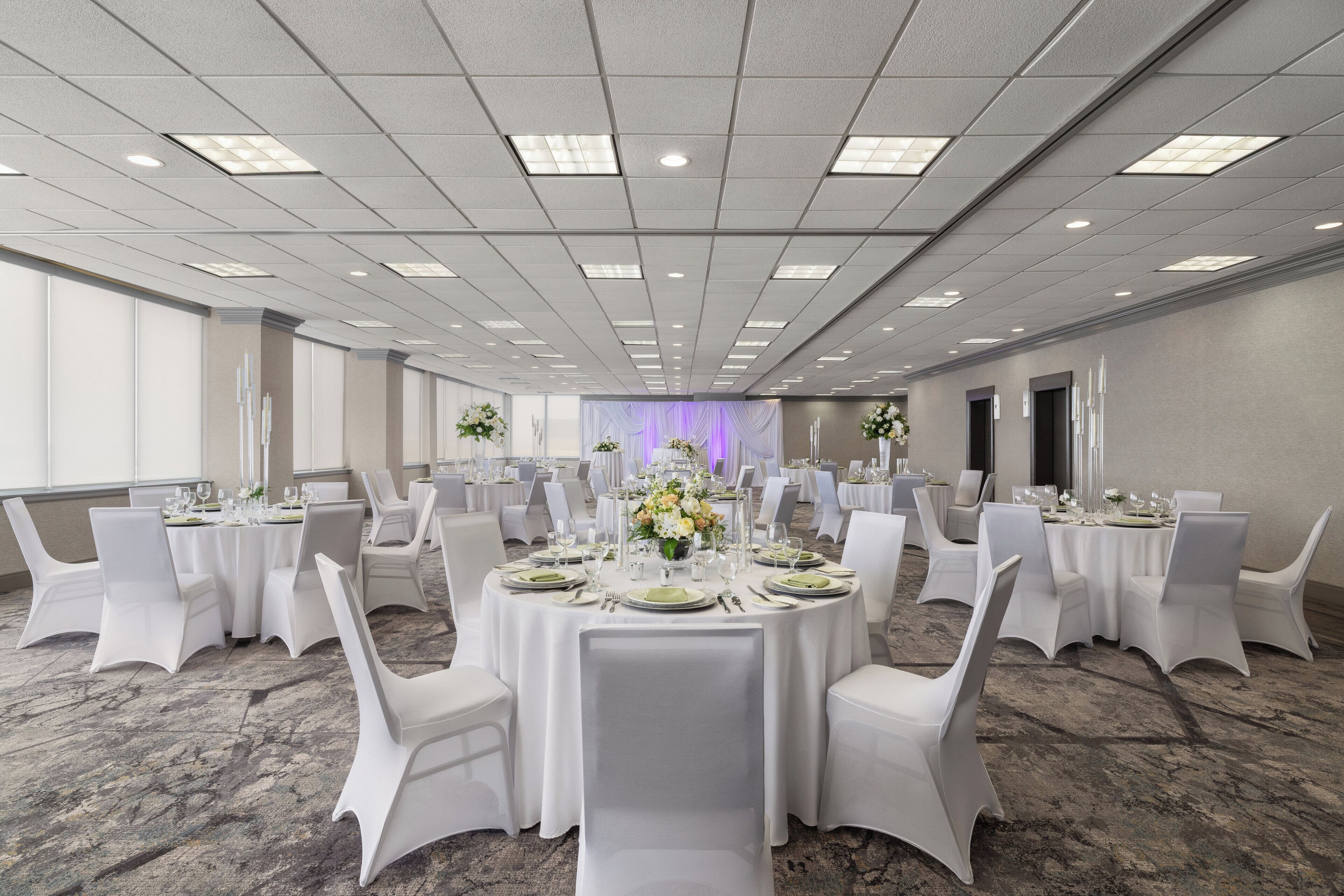Un salón de conferencias con mesas redondas, sillas blancas y centros de mesa con flores.