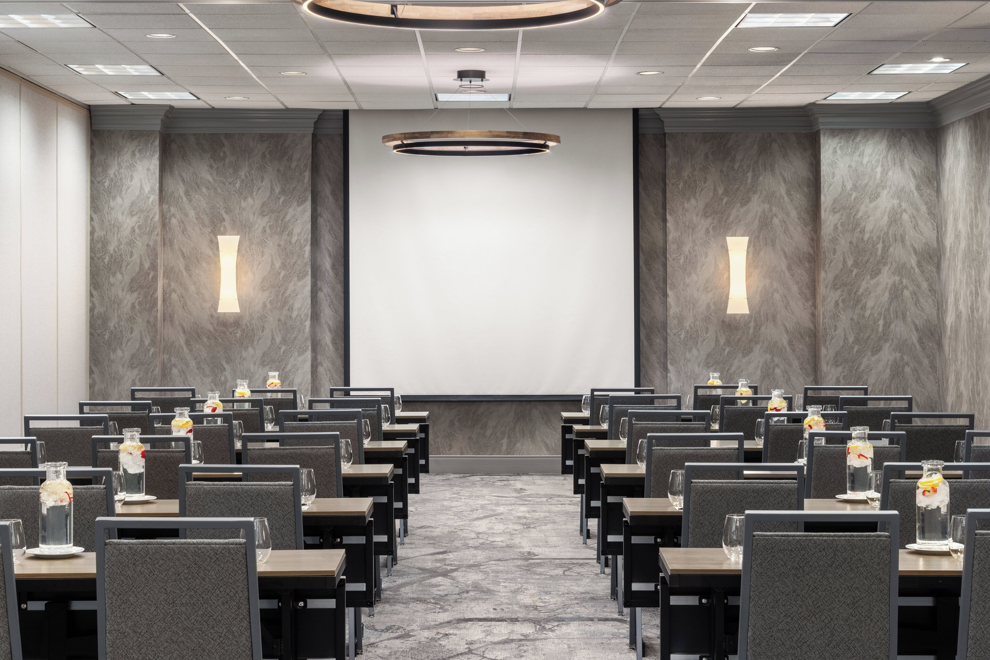 A conference room with rows of tables and chairs, equipped with water bottles and glasses, and a large screen at the front.