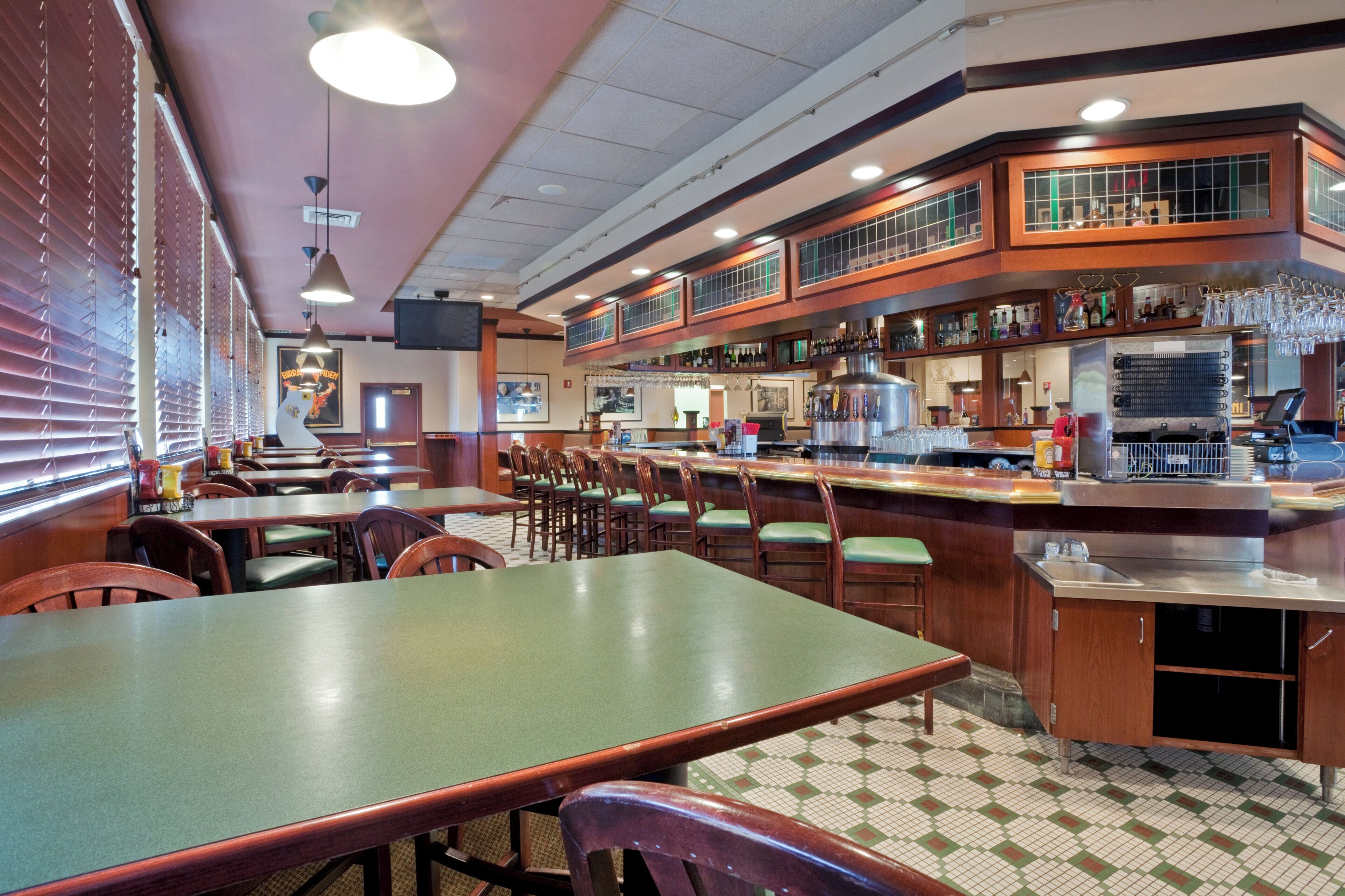 Un restaurante con varias mesas y sillas, una zona de bar con grifo y vasos, y suelo de baldosas.