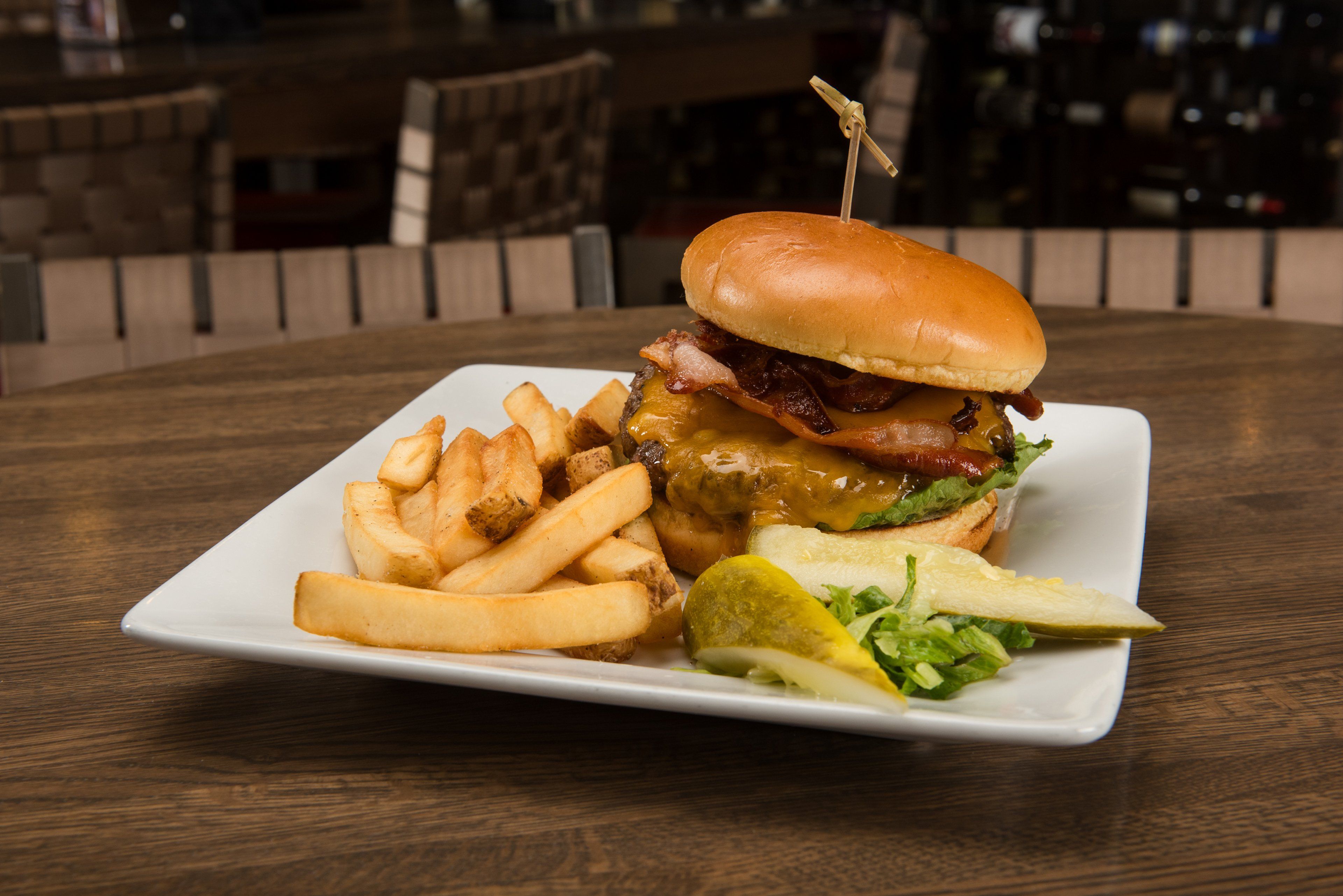 Une assiette carrée contenant un hamburger, des frites et un cornichon, posée sur une table en bois.
