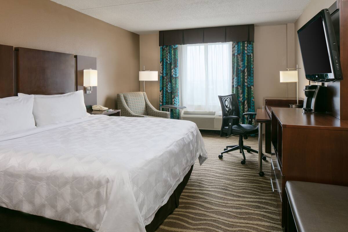 A hotel room with a large bed, two chairs, a desk with a computer, and a TV.