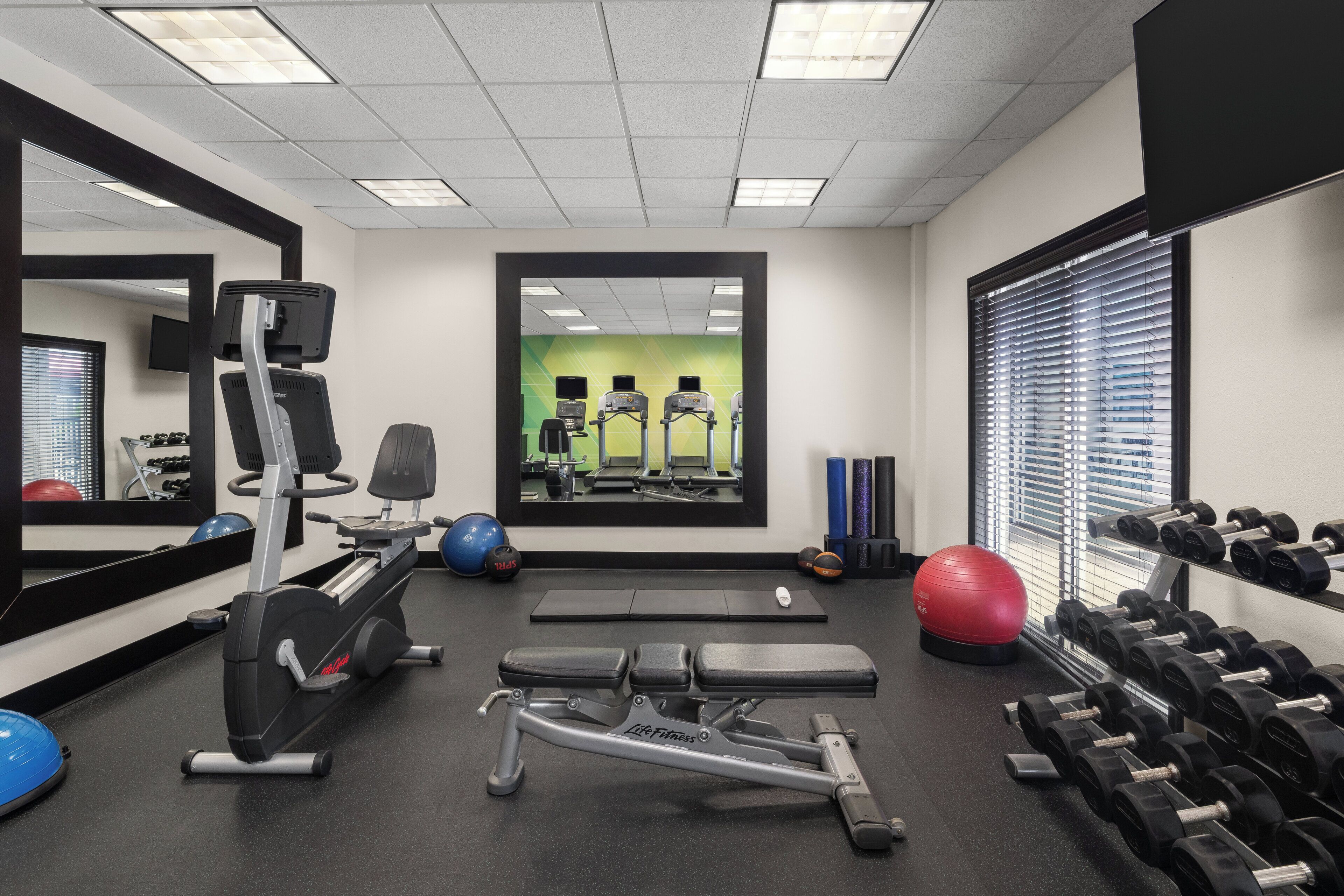 A gym with exercise equipment, a mirror, and a window with blinds.