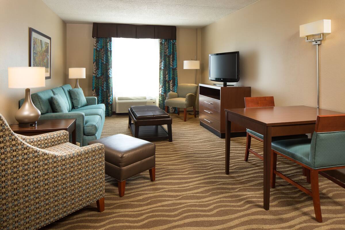 A hotel room with a sofa, armchair, ottoman, TV, and a dining table.