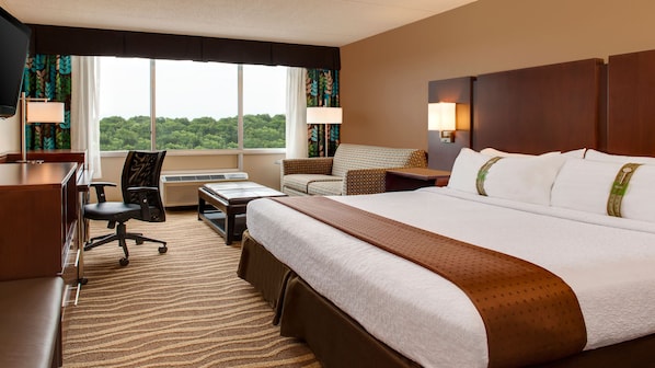 A hotel room with a large bed, a desk, a chair, and a sofa. There is a window with curtains and a view of greenery outside.
