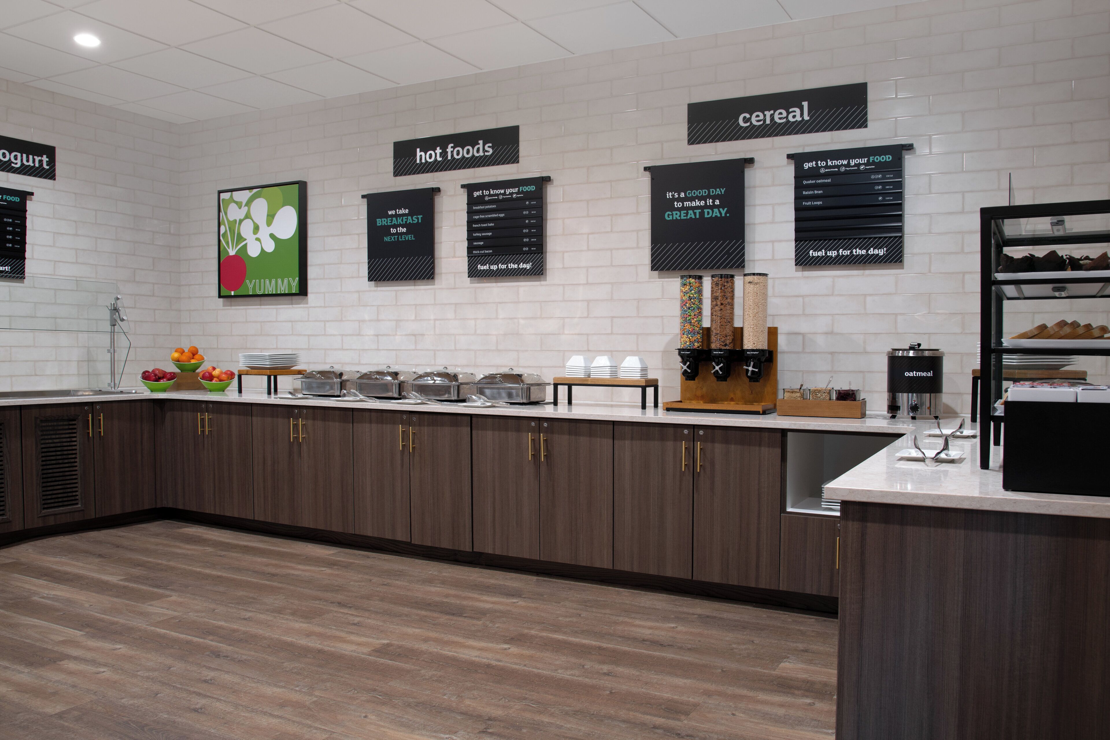 A modern breakfast area with a counter displaying various food items and a wall with labeled sections for hot foods and cereal.