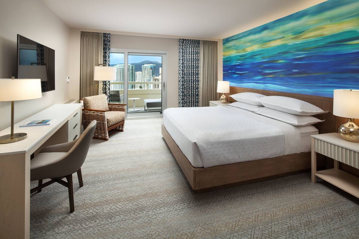 A hotel room with a large bed, a desk, a chair, a TV, and a painting on the wall.