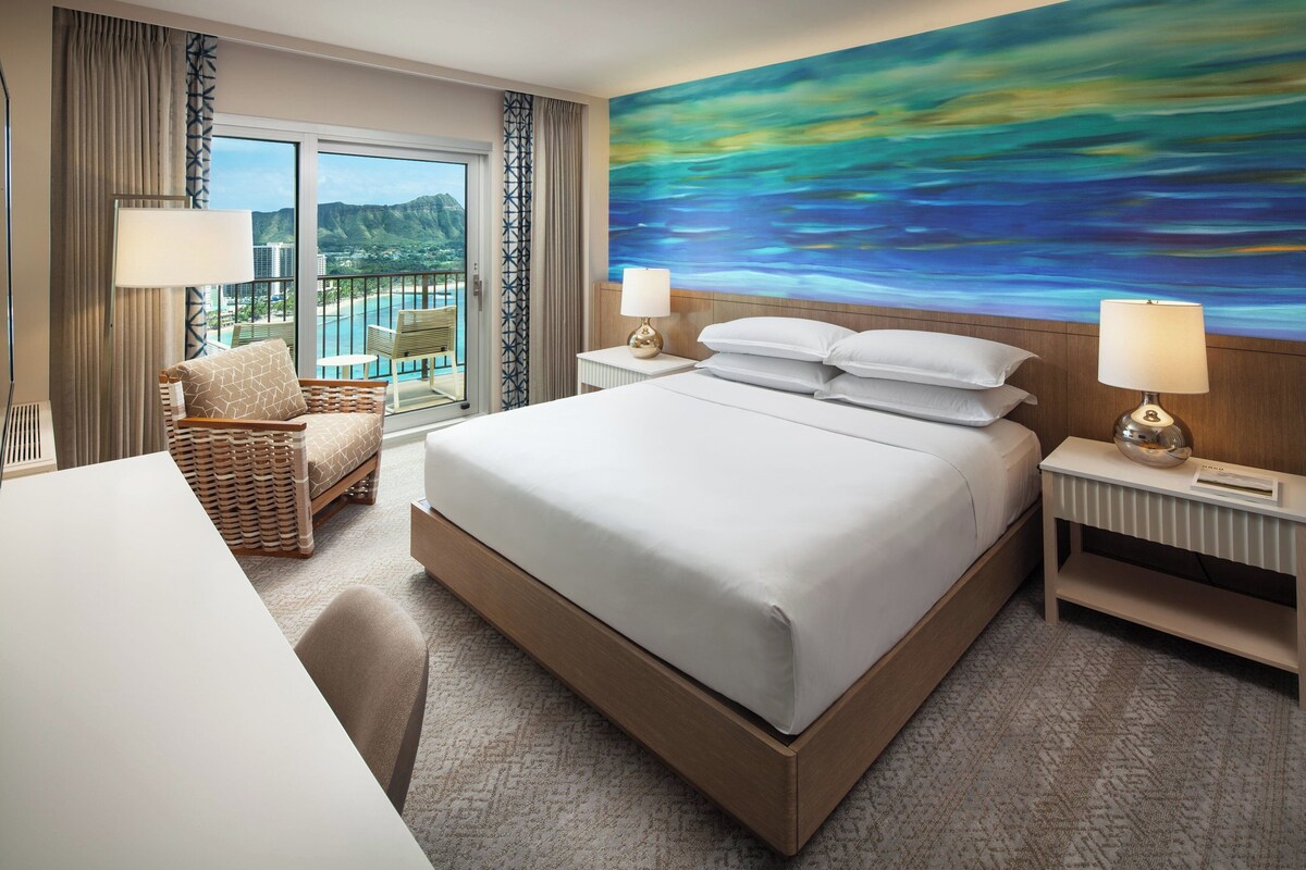 A hotel room with a large bed, two bedside tables with lamps, a chair, and a view of a scenic landscape through a large window.