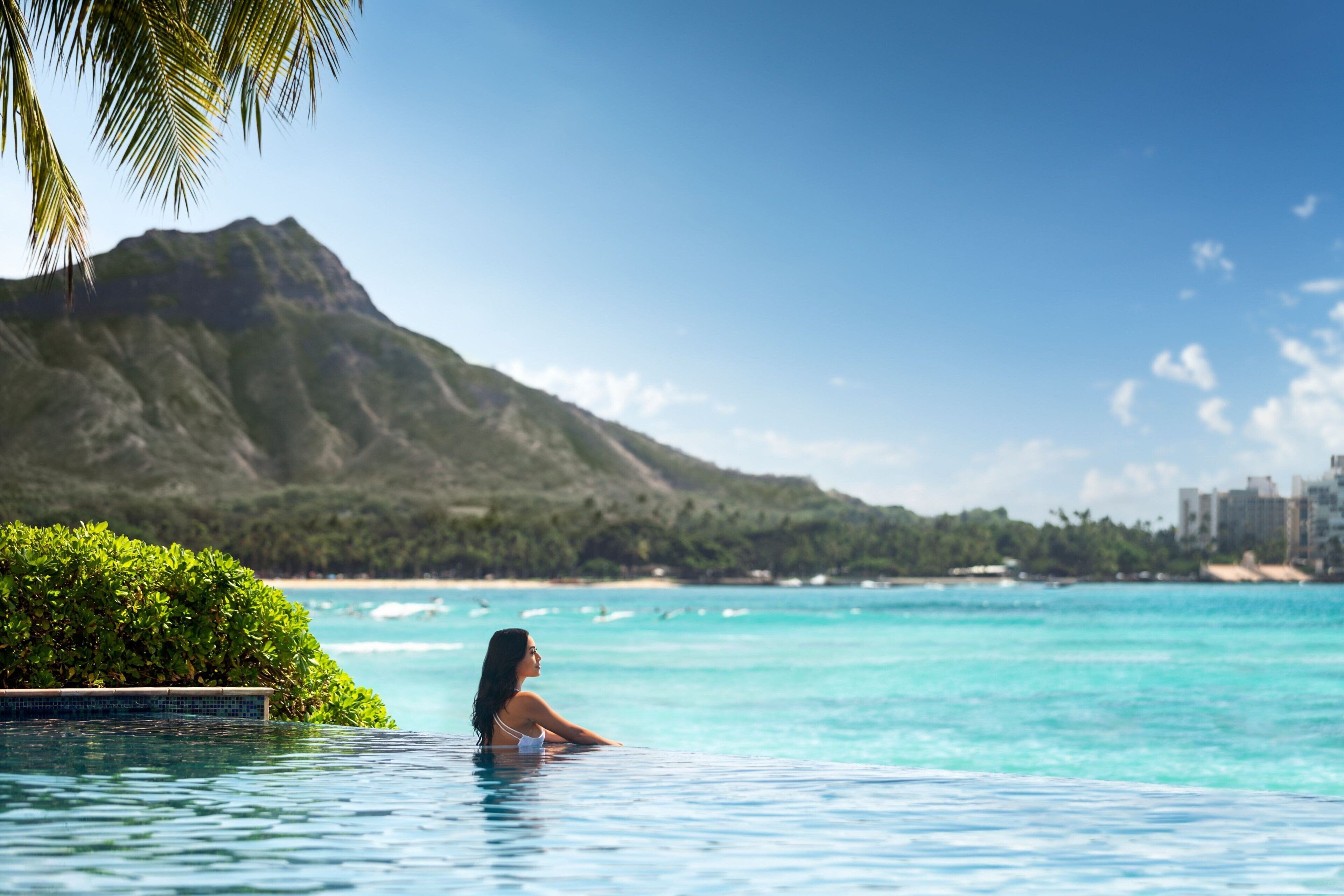 A woman in a swimming pool with a mountain and beach in the background.