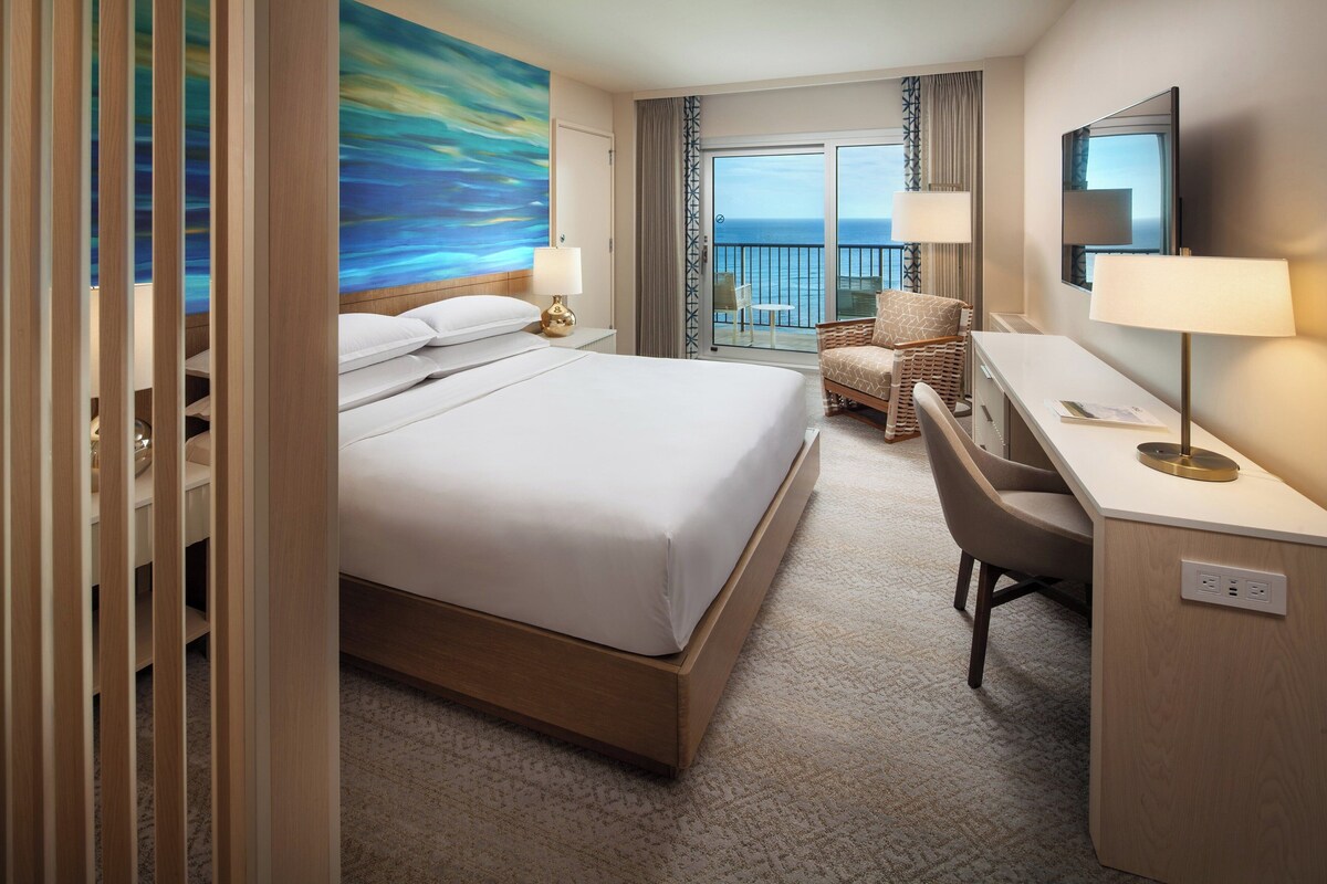 A hotel room with a large bed, a desk, a chair, a TV, and a balcony with a view of the sea.