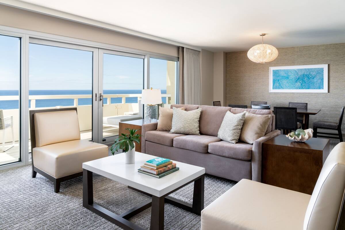 A modern hotel room with a balcony, a sofa, a coffee table, and a dining area.