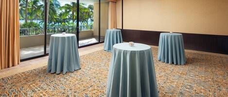 Un salón de conferencias con tres mesas redondas, una alfombra estampada y un amplio ventanal con vistas a palmeras.