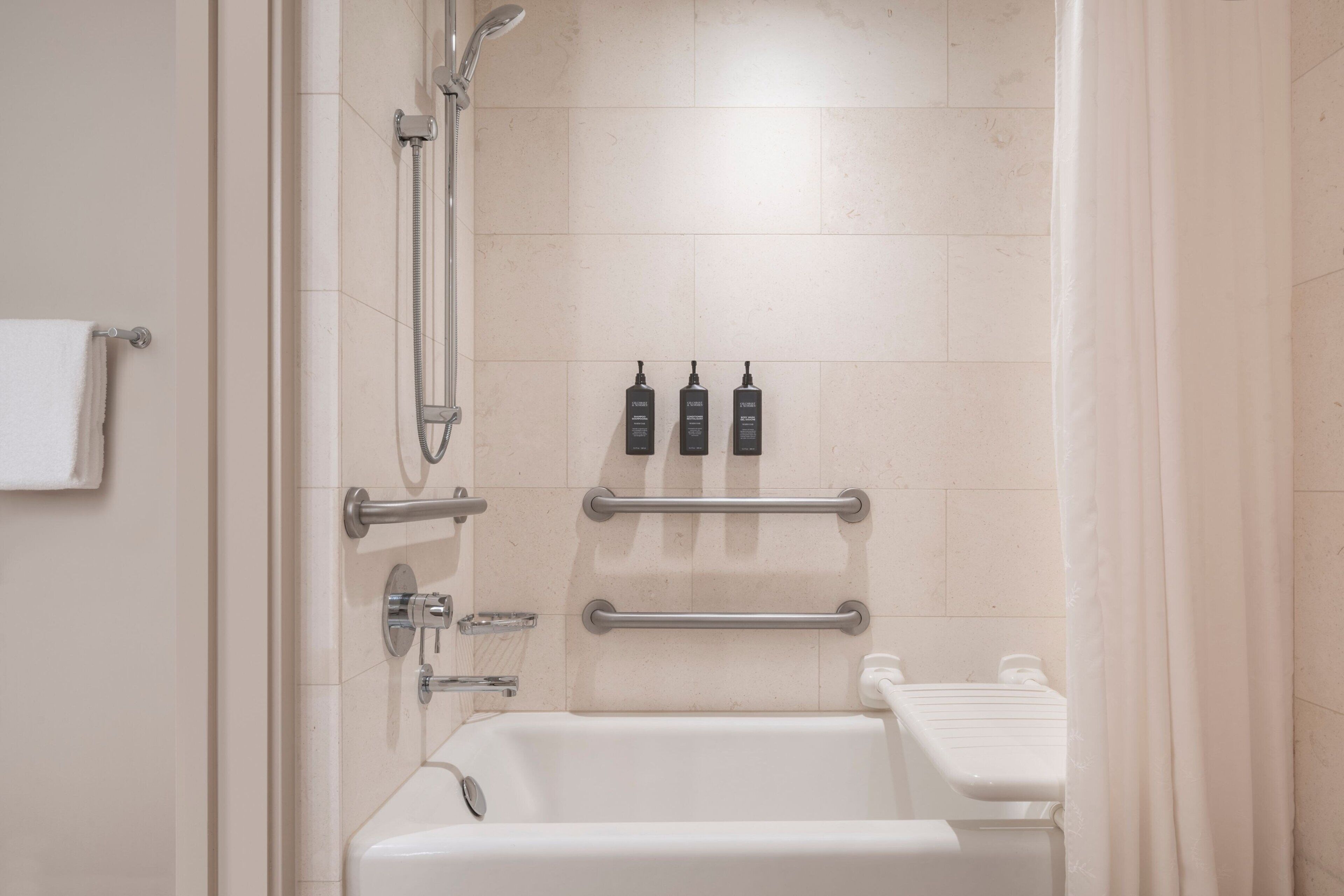 Baño con bañera, ducha y barras de apoyo. Hay tres botellas de champú en la pared.