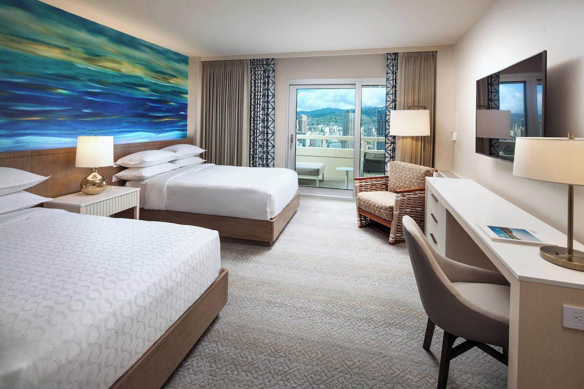 A hotel room with two beds, a desk, a chair, a large painting, and a view of the city through the balcony doors.