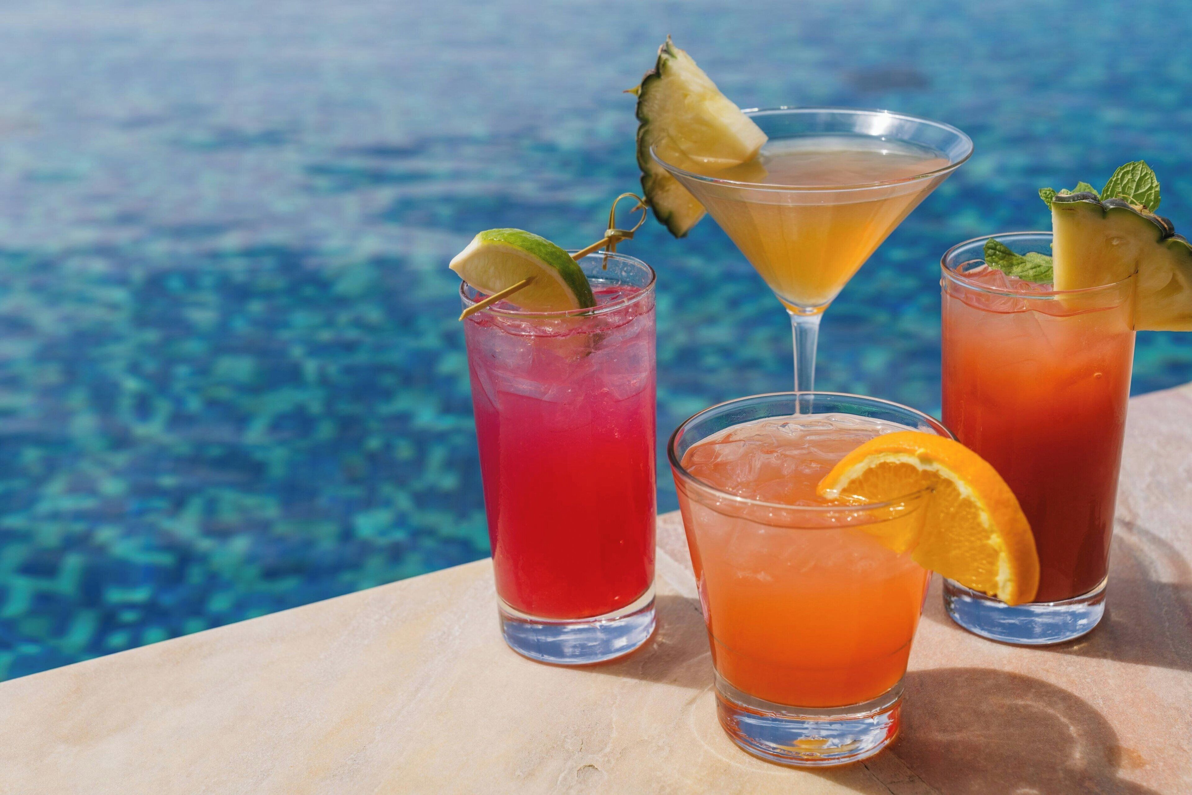 Three drinks with fruit garnishes on a table by a pool.