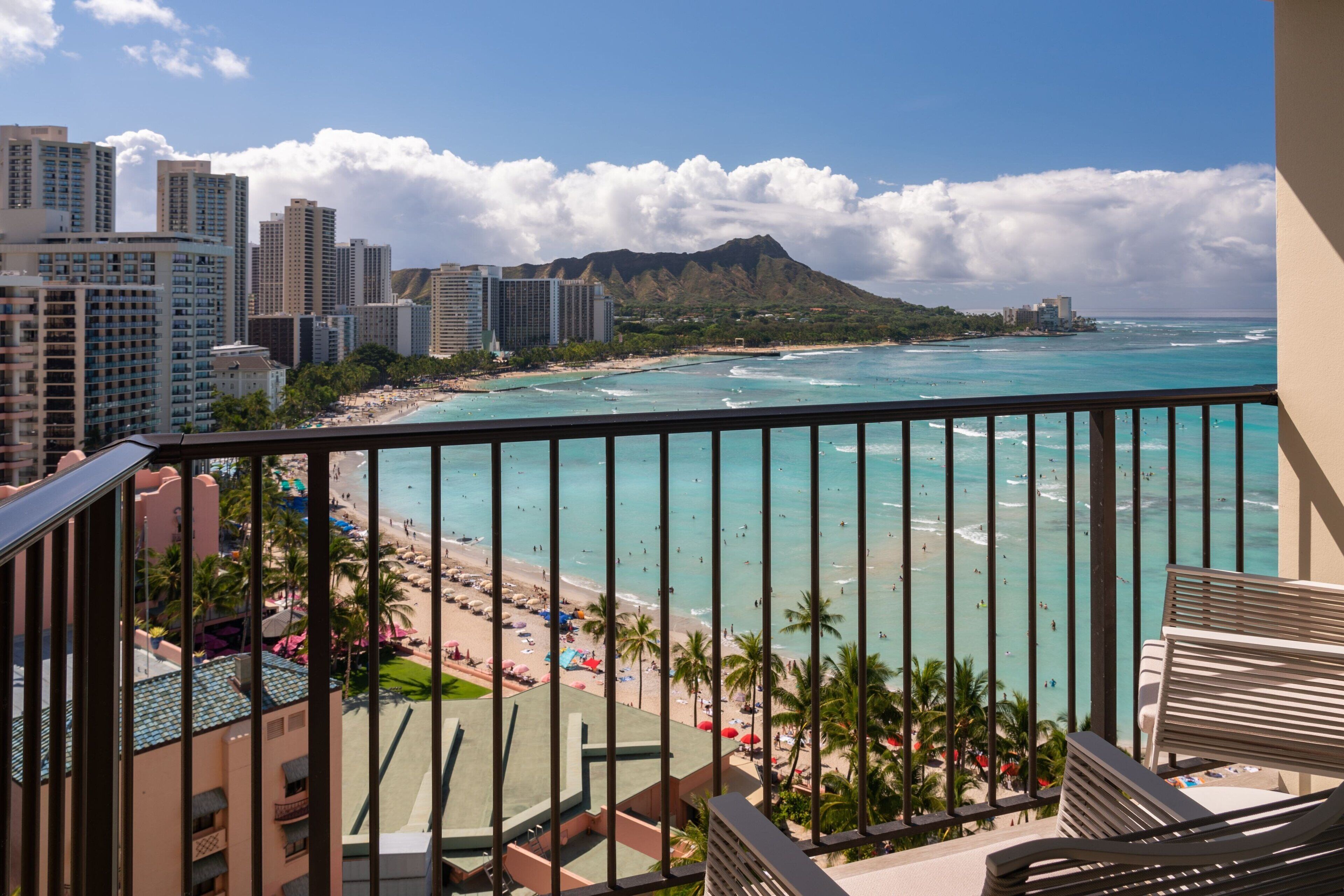 A balcony with a view of a beach and ocean, with buildings in the background.