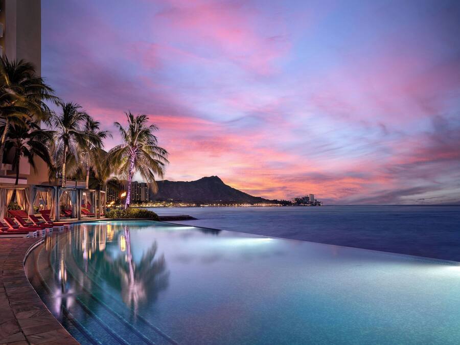Vista a la piscina con sillas de descanso, palmeras y una puesta de sol sobre el océano.