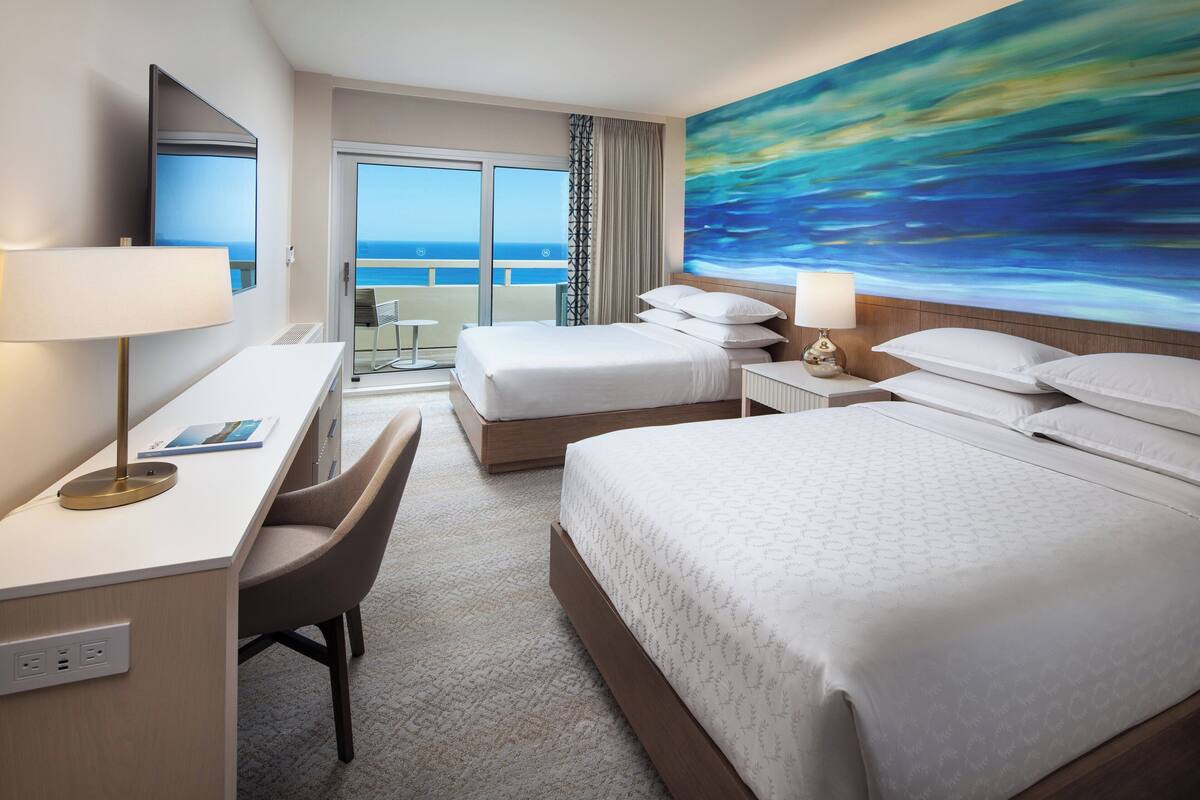 Habitación de hotel con una cama grande, un escritorio con silla, un televisor y un cuadro con vista al océano azul.