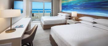 A hotel room with a large bed, a desk with a chair, a TV, and a painting of a blue ocean view.
