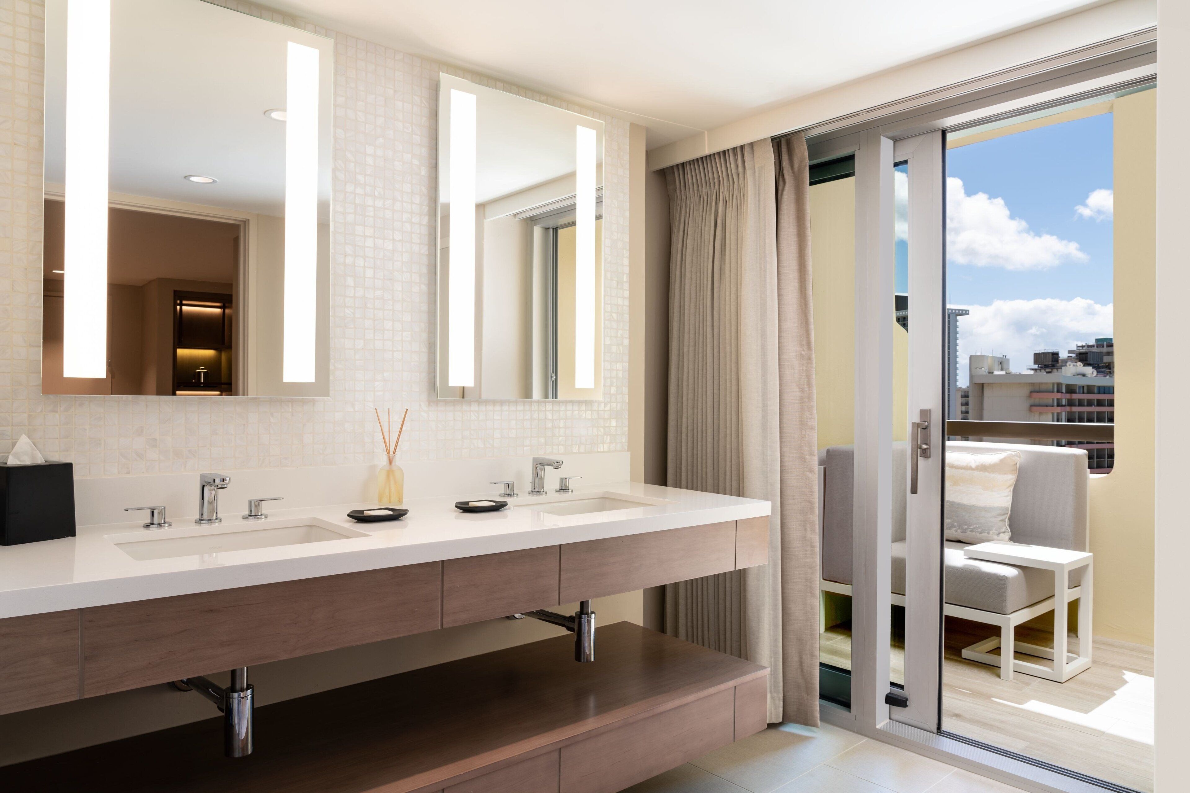 A modern bathroom with a dual sink vanity, a large mirror, and a sliding glass door leading to a balcony with outdoor seating.
