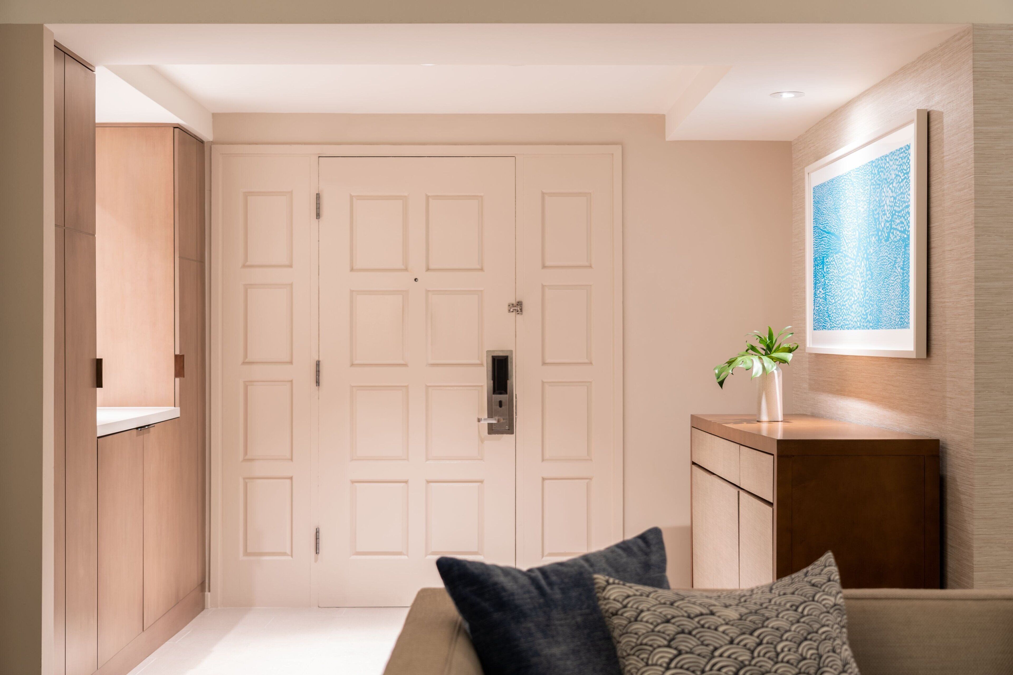 A modern hotel room with a wooden wardrobe, a white door, a small bedside table with a plant, and a patterned pillow.