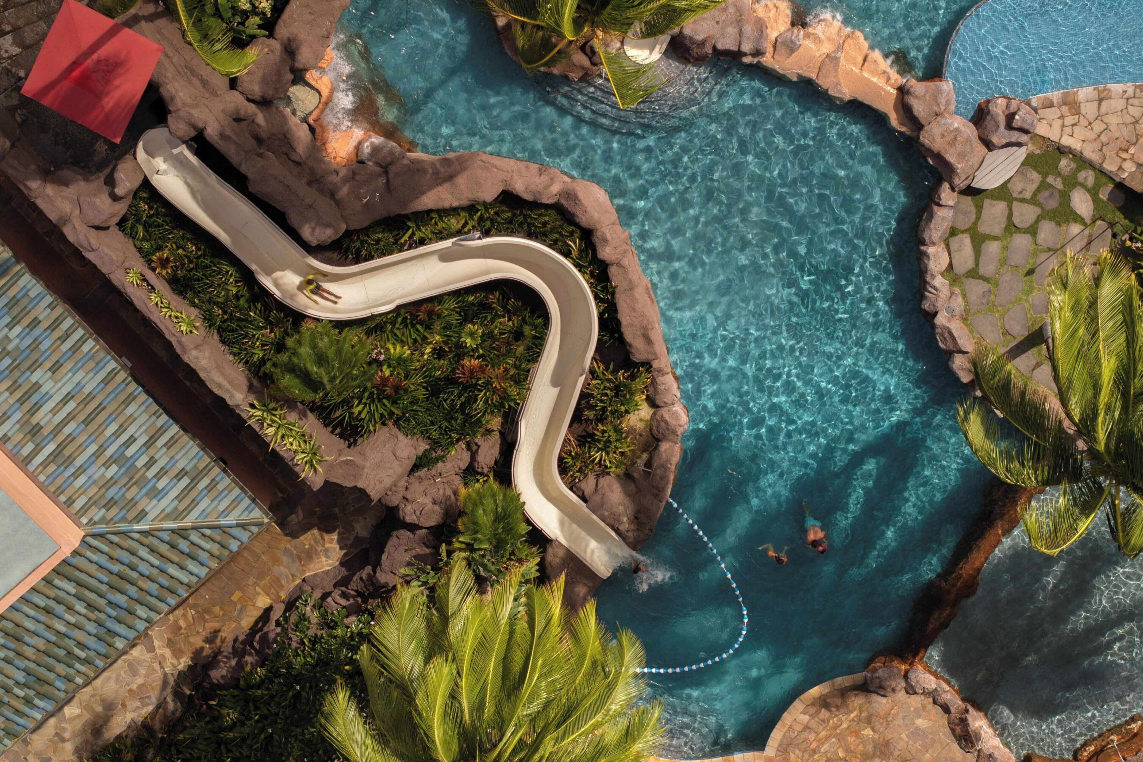 A swimming pool with a winding water slide, surrounded by rocks and tropical plants.