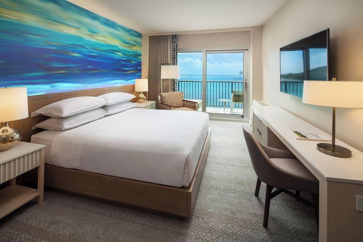 Habitación de hotel con una cama grande, un escritorio, una silla, televisión y balcón con vista al océano.