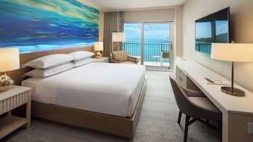 A hotel room with a large bed, a desk, a chair, a TV, and a balcony with a view of the ocean.
