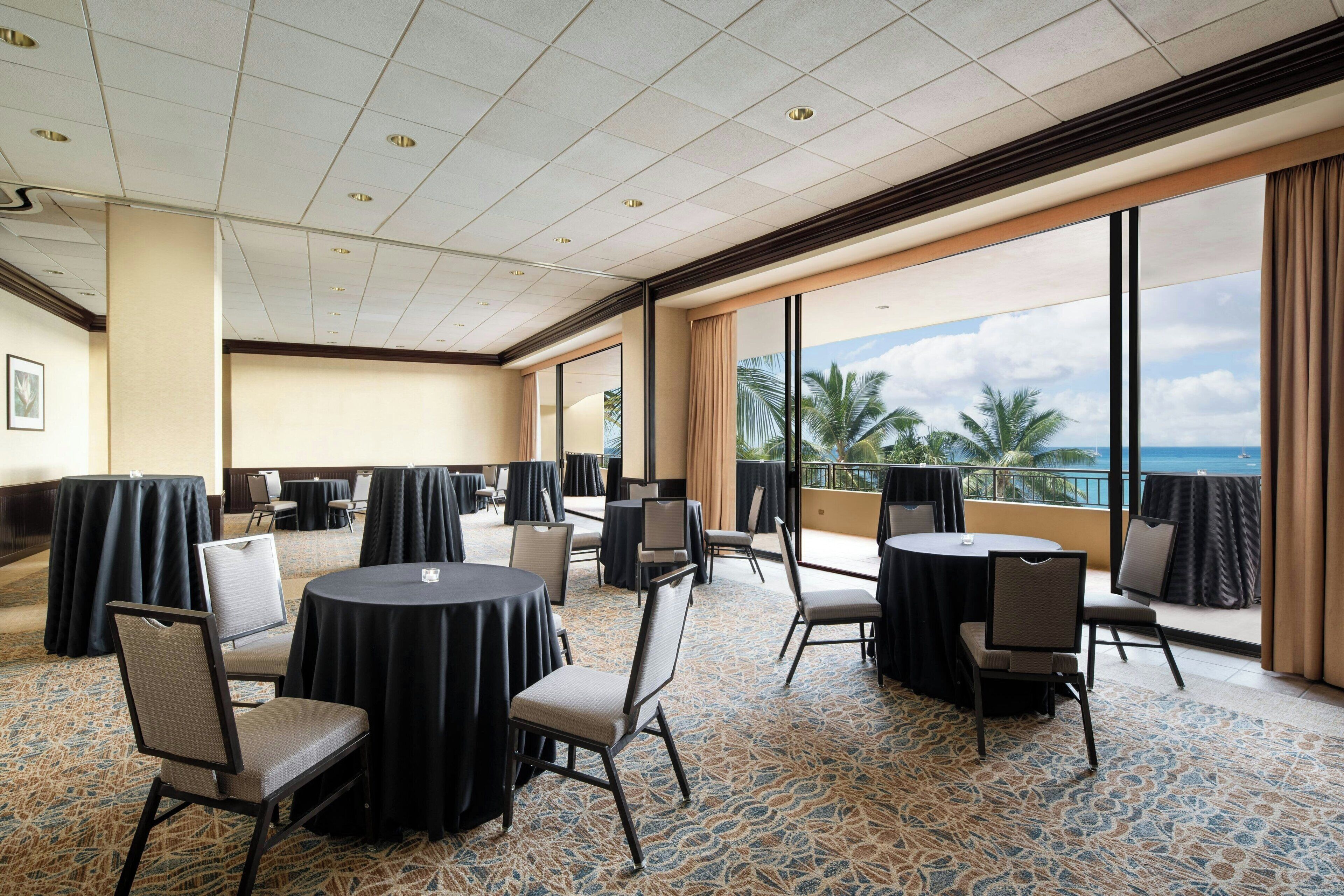 A conference room with round tables and chairs, large windows with a view of palm trees and the ocean, and a patterned carpet.
