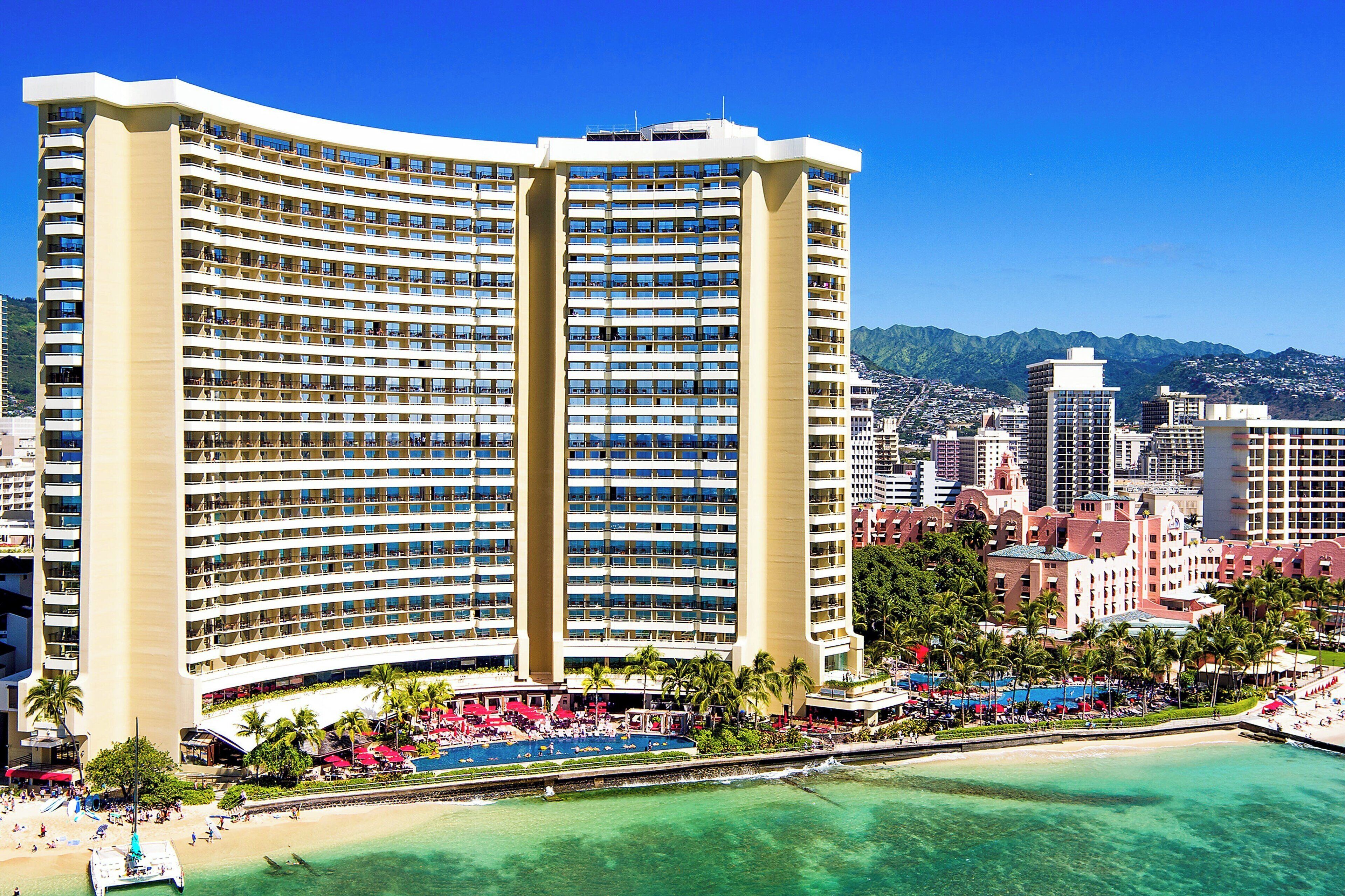 A large hotel with multiple floors and balconies, a beachfront location, and a pool area with umbrellas.