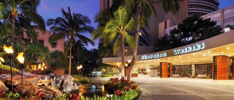 Entrada del hotel Sheraton Waikiki con un jardín ajardinado, palmeras y una fuente de agua.