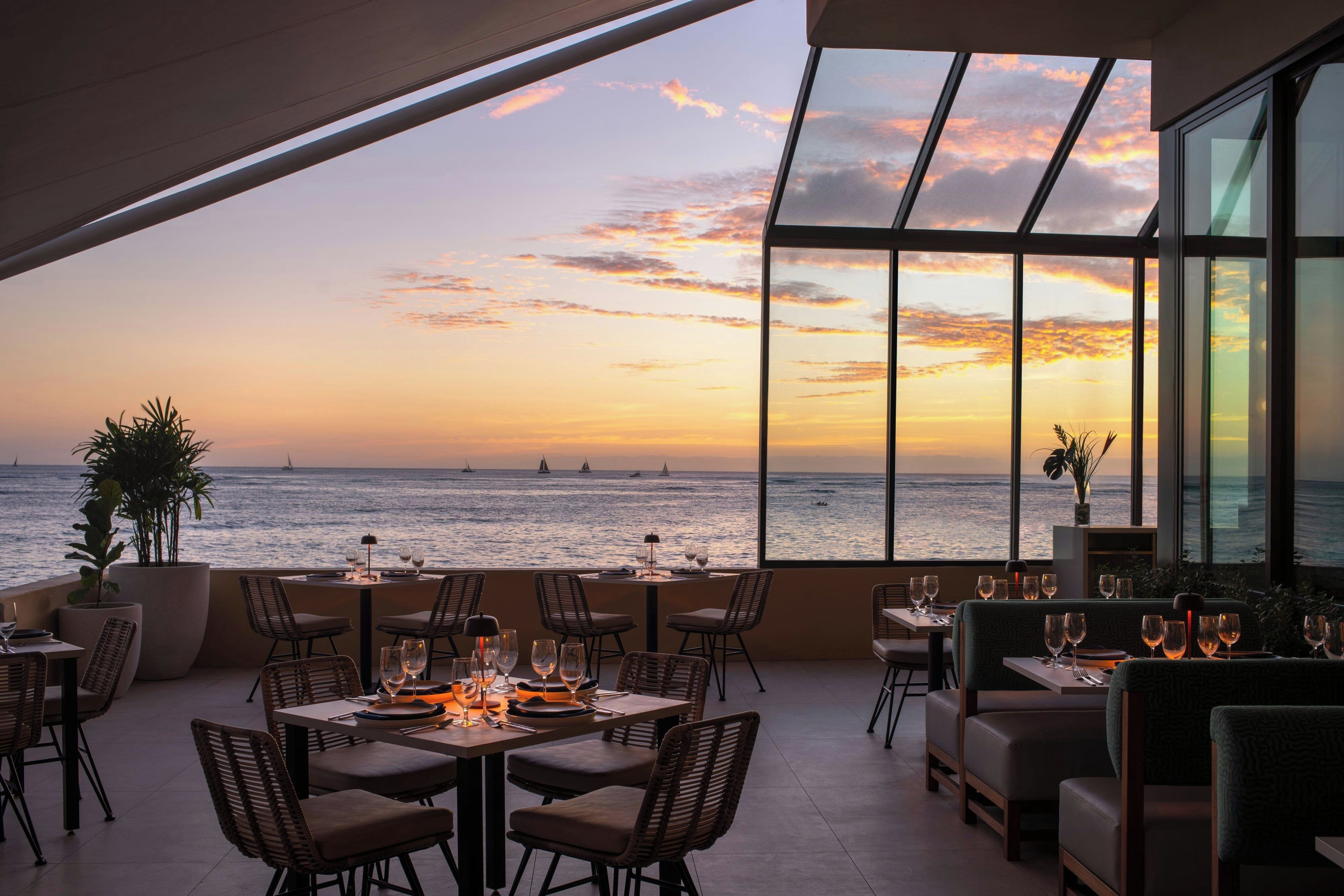 Restaurante com mesas e cadeiras voltadas para um pôr do sol sobre o mar.