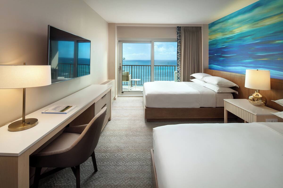 Habitación de hotel con una cama grande, un escritorio con una silla, un televisor y un balcón con vista al mar.