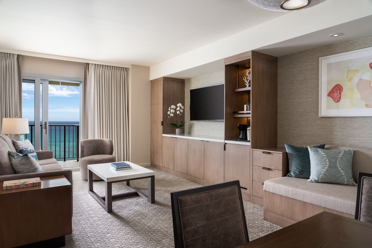 A modern hotel room with a balcony, a flat-screen TV, a sofa, a coffee table, and a seating area with cushions.